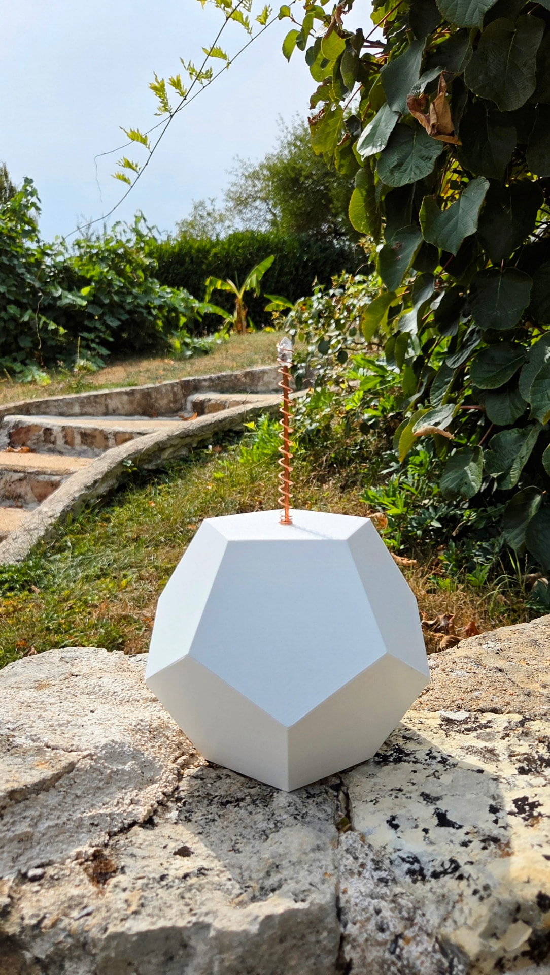 Bio-harmonisateur Ethérion Kairos : dodécaèdre blanc mat surmonté d'une antenne de cuivre en spirale et d'un diamant de Herkimer, posé sur un mur de pierre dans un jardin